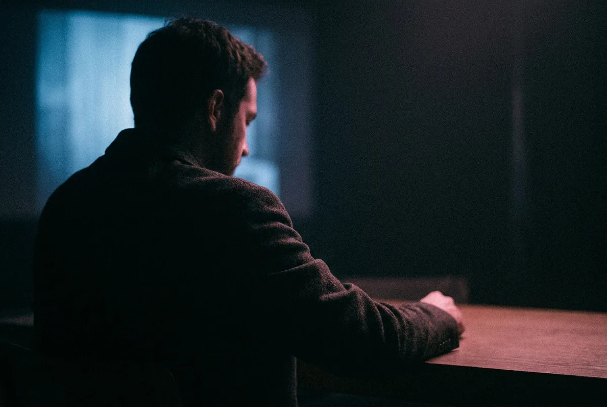 A figure alone in low light, turned away, reading by the glow of a screen